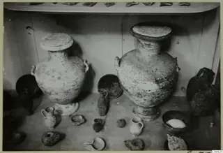 Two large two-handled jars with lids, with smaller vessels and lamps, displayed on a shelf