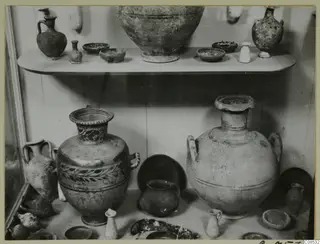 Pottery vessels displayed on two shelves in a glass case, including large two-handled jars and small juglets.