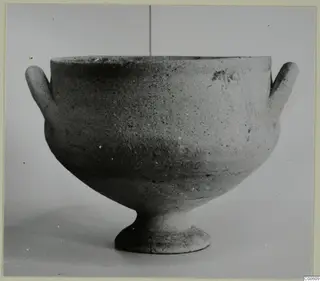 Unpainted pottery bowl with two small horizontal handles and a short footed base, photographed against a plain grey background.