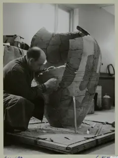 Man restoring a large broken pithos-type jar on a wooden board in a workroom