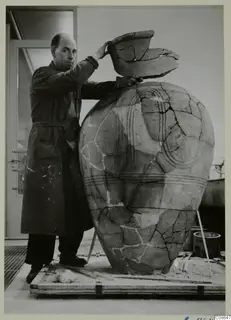 Man in a dark work coat with one hand on a broken rim fragment atop a large mended pottery vessel