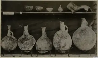 Pottery vessels displayed on a shelf: three bowls and a small juglet above, five globular juglets on wire stands below, from Lapithos.