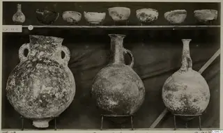 Pottery jars, a jug, a flask and a row of bowls displayed on a shelf against a dark cloth, from Lapithos Markides