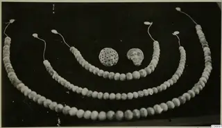 Three strands of pale beads with a perforated round pendant and a small face-shaped pendant on a dark background