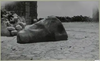 Large rounded stone block lying on a paved ground beside a stone wall, with cacti behind, at Paphos area.