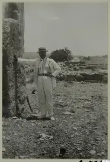 Man in a light suit and boater hat standing beside a large stone block, with low stone walls behind him