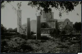 Stone building with a tall bell tower and two standing pillars in front of it, with tree branches across the top
