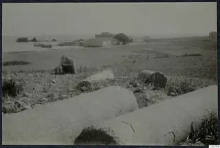 Fallen column drums in the foreground, with small boats on the water and low buildings along the shore beyond.