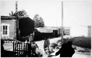 Woman in a hat, a man and a donkey beside an iron gate on a village street.