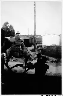 Woman in a hat and long coat riding a camel past low buildings and a wooden pole