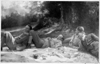 Four men lying and reclining on a blanket under trees, one of them smoking a pipe.