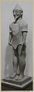 Terracotta statue of a standing male figure with a pointed cap and beard, on a rectangular wooden base