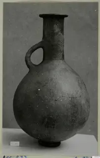 Red Polished III jug with a single loop handle and flared rim, photographed on a white plinth against a grey backdrop