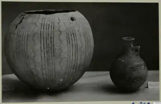 Two painted pottery vessels, a large globular jar and a small juglet, standing on a pale surface against a dark backdrop