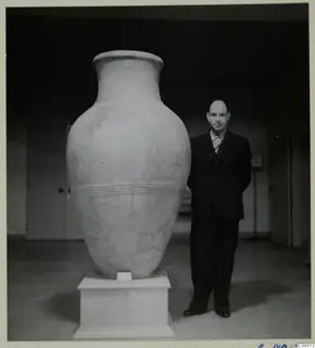 Large pale pottery pithos on a white plinth, with a man in a dark suit standing beside it for scale.