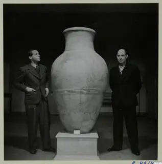 Two men stand beside a very large pottery pithos ware jar on a white pedestal in a dark room.