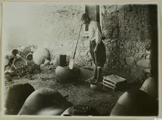 Potter standing in a courtyard at Klirou beside a bowl of clay, with large jars and a stone and mud-brick wall behind him