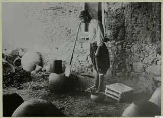 Potter treading clay in a courtyard at Klirou, with large storage jars and a spade beside a stone and mud-brick wall