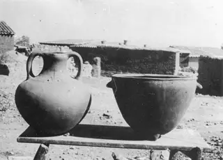 Two new pottery vessels, a two-handled jug and a wide basin, on a wooden table in a village yard with mud-brick houses behind