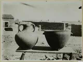 Two new clay pots on a wooden table in a village yard, with mud-brick houses behind.