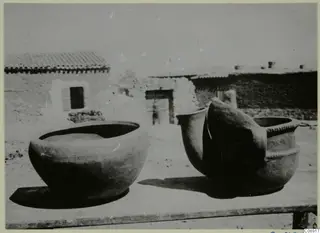 Two large clay pots on a wooden table in a courtyard at Klirou, with low buildings behind