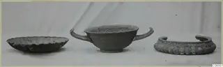 Three corroded silver objects on a pale surface: a shallow scalloped dish, a two-handled cup and a wide ring-shaped object.