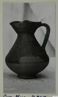 Dark jug with a pinched mouth and a single strap handle, photographed on a plain surface against a grey backdrop