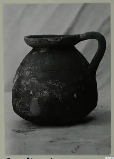 Dark handled jug with a wide flat rim and faint incised lines on the body, photographed against a grey backdrop
