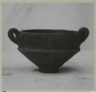 Dark grey two-handled bowl with a wide rim, a ridge at the shoulder and a short footed base, photographed on a plain surface.