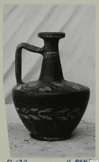 Dark-surfaced juglet with a single strap handle and pale painted decoration, photographed on a mounted card