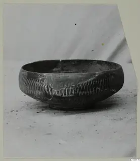 Dark bowl with bands of pale incised strokes on its body, shown on a plain surface against a light backdrop