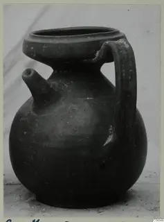 Dark globular jug with a side spout and a strap handle, shown on a pale surface
