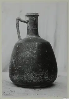 Dark jug with a single loop handle, narrow neck and flaring rim, photographed against a pale backdrop