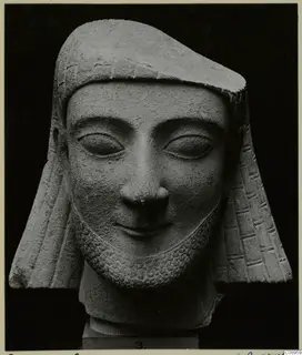Limestone head of a bearded figure in a close-fitting headdress with side flaps, set on a numbered base