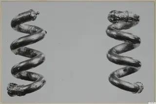 Two gold spiral objects, each a coil of tubular wire with decorated ends, shown side by side.