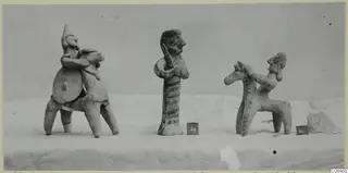 Three terracotta figurines on a pale cloth: two riders on four-legged forms and a standing figure with a striped body.