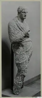 Full-length statue of a standing figure in rough, pitted limestone, with a draped cloak and arms folded at the chest