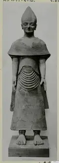 Terracotta statue of a standing figure with a tall conical headdress, a ridged cape and a long robe