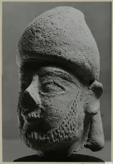Terracotta head of a bearded figure wearing a tall rounded cap, shown against a plain grey backdrop