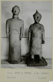 Two standing clay statues of human figures, one on a dark wooden base, against a pale draped backdrop