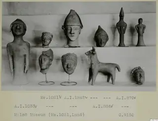 Ten small clay figurines and heads arranged on two shelves, including a standing figure, a capped head and a quadruped figure