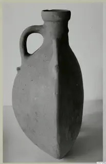Pottery flask with a loop handle and a flattened body, standing on a pale surface against a grey backdrop.