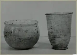 Two blown glass vessels: a rounded bowl on the left and a ribbed beaker on the right, on a plain surface