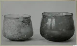 Two blown glass vessels standing side by side on a pale surface, one wider with a flared rim, one rounder and deeper.