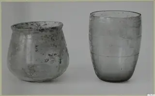 Two blown glass vessels side by side on a pale background: a squat, weathered jar left and a tall beaker with ridges right.