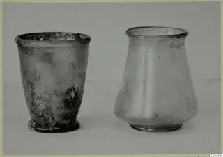 Two blown glass vessels, a beaker and a jar, standing side by side on a pale surface