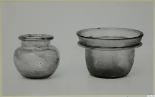 Two small glass vessels: a round-bodied jar on the left and a wide cup-like pyxis with a flanged rim on the right.