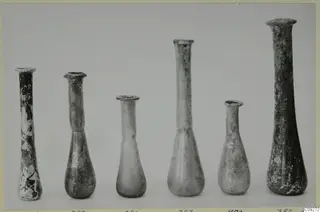 Six blown glass flasks and unguentaria of differing heights standing in a row against a grey background
