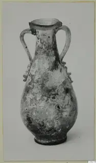 Blown glass amphoriskos with two handles and a flared rim, its surface weathered, shown against a plain grey backdrop