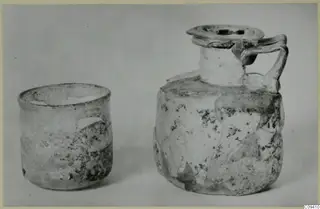 Two weathered glass vessels on a grey backdrop: a straight-sided beaker and a jug with a strap handle and disc-shaped mouth